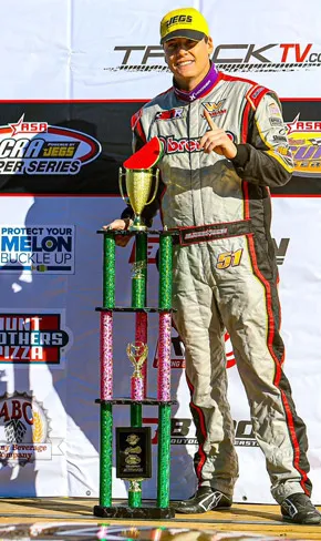 A Cordele Motor Speedway winner holding the victory-lane trophy in front of the Protect Your Melon — Buckle Up sponsor banner